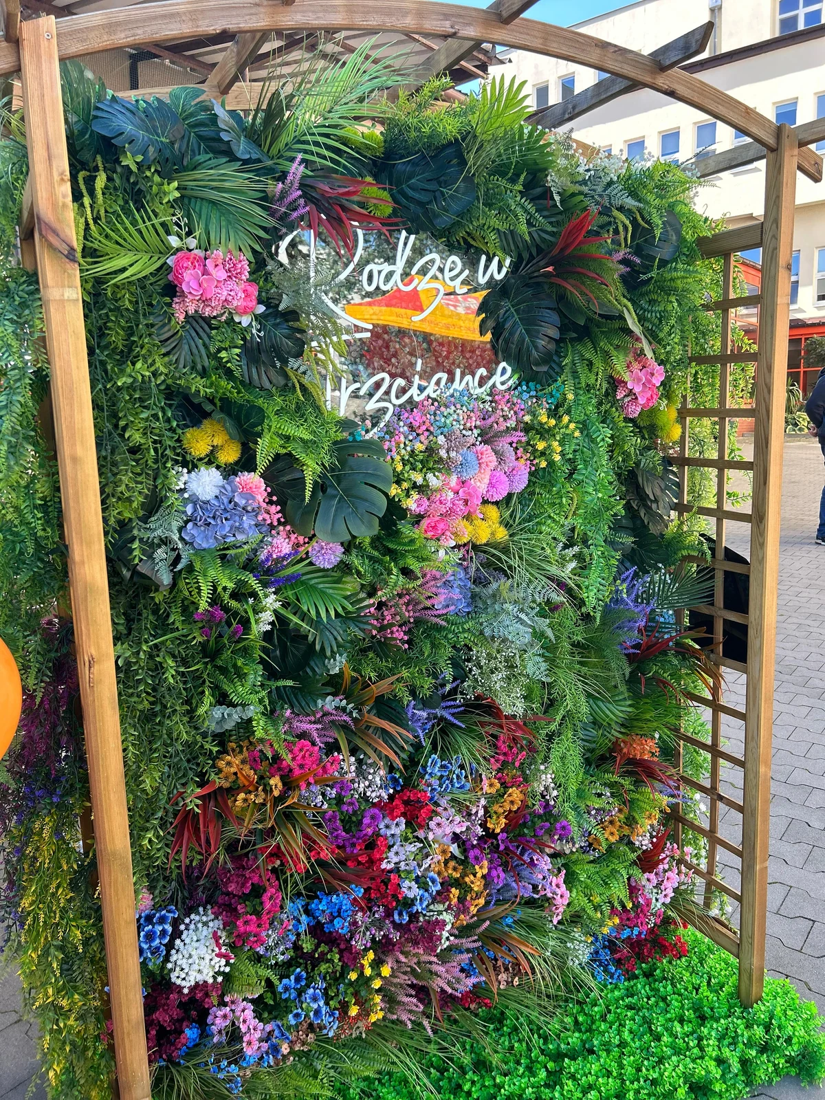 Prawdziwa fotościanka Green Thumbs na pikniku w szpitalu w Trzciance: drewniana rama, tropikalne liście, kolorowe kwiaty i napis Rodzę w Trzciance.